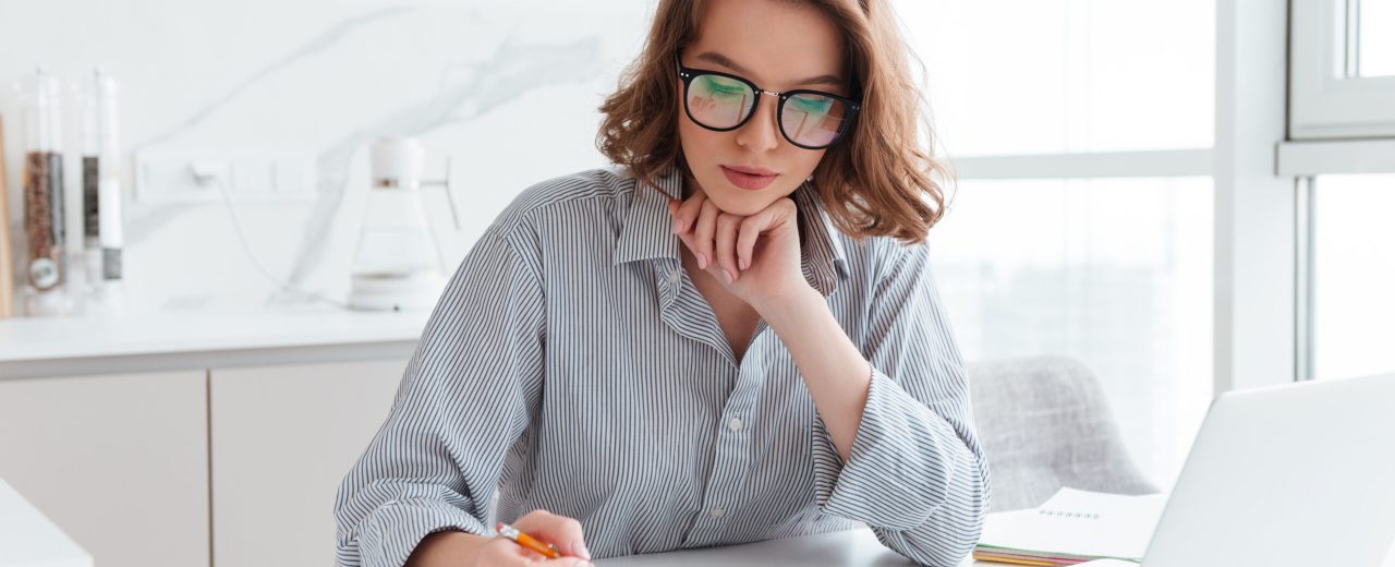 Young concentrated businesswoman in glasses and striped shirt working with papers at home
