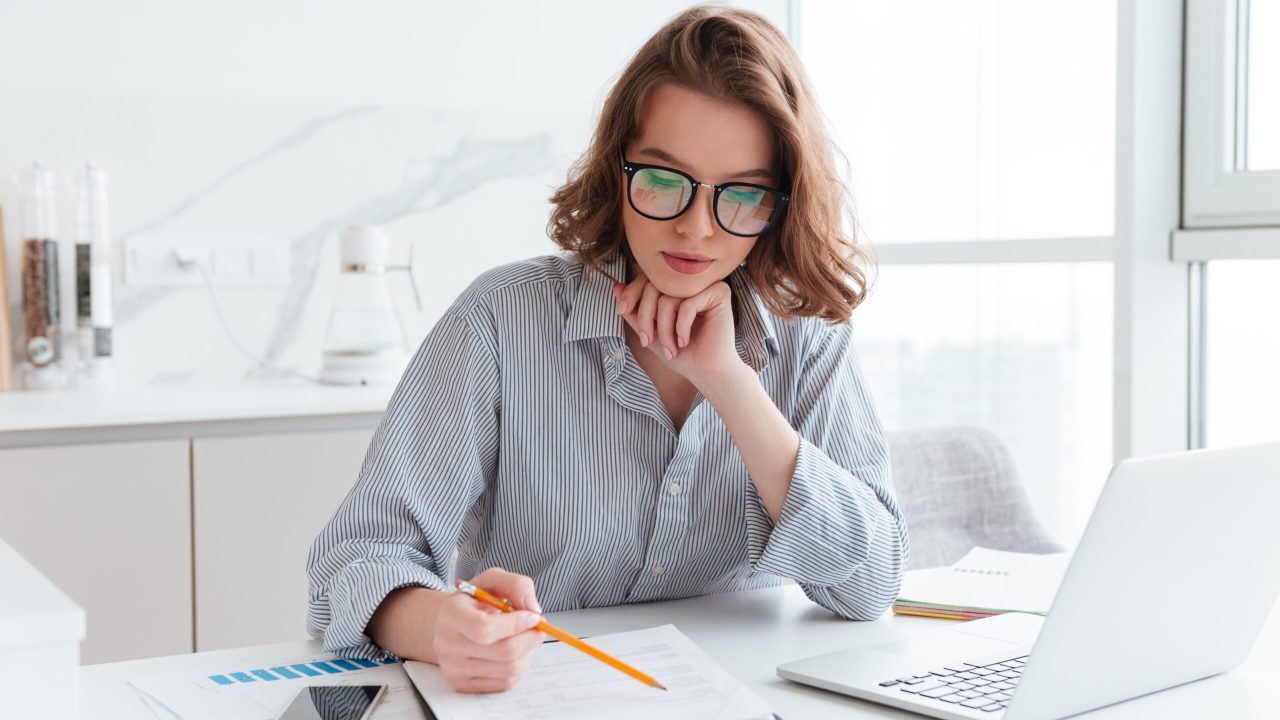 Young concentrated businesswoman in glasses and striped shirt working with papers at home
