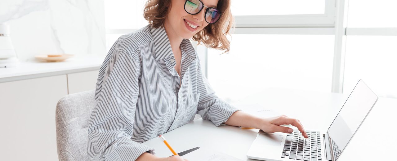 Young attractive smiling girl in glasses and striped shirt working with documents and computer while siting at table in light kitchen