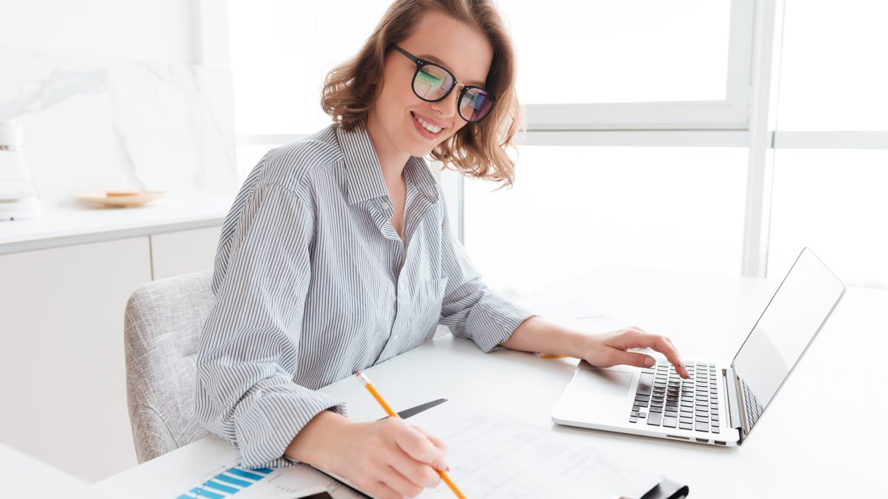 Young attractive smiling girl in glasses and striped shirt working with documents and computer while siting at table in light kitchen